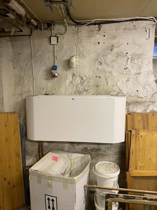 Källare med ojämn betongvägg och synliga rör, elektriskt skåp och vit radiator ovanför förpackningar och byggmaterial.