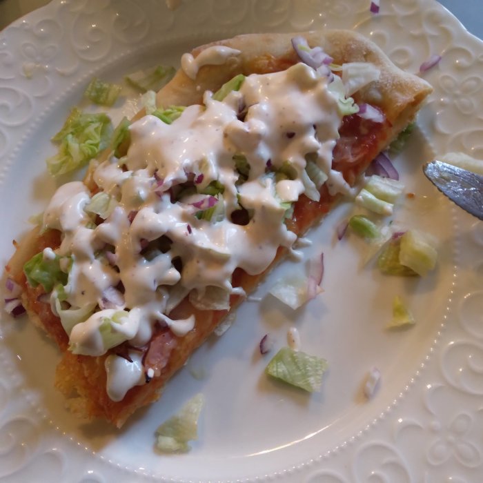 Bit av hemgjord pizza med krispig botten, tomatsås, ost, sallad och vit sås på ett vitt fat.