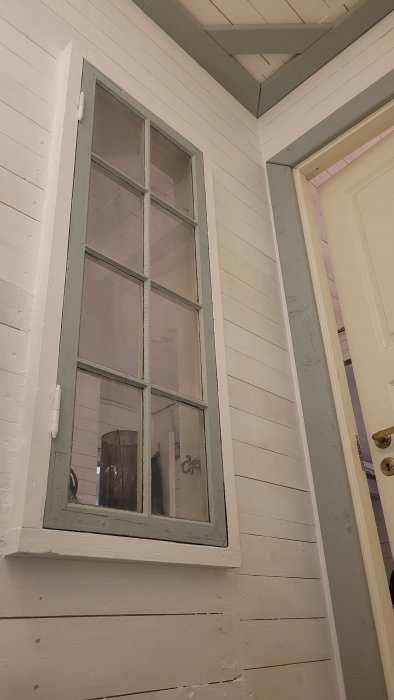 Ett nyrenoverat trapphus med målade vita väggar och en fönster med grå karm intill en stängd dörr, renoveringsarbete pågår.