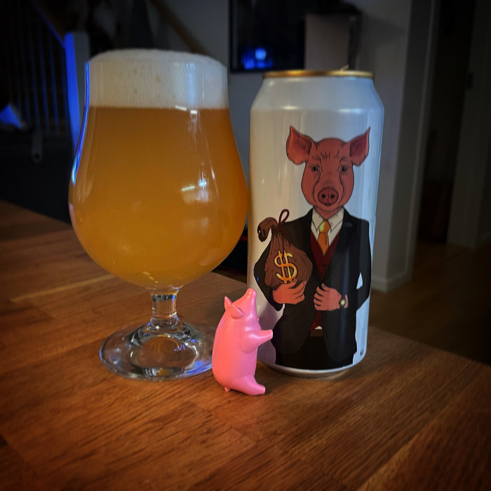 Ölglas fyllt med öl bredvid en ölburk med tecknad gris i kostym och en liten plastgris framför.