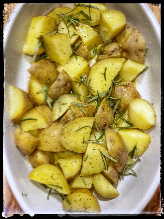 Krossad kokt potatis kryddad med rosmarin, flingsalt och olivolja i en ugnsform, redo för att gräddas.