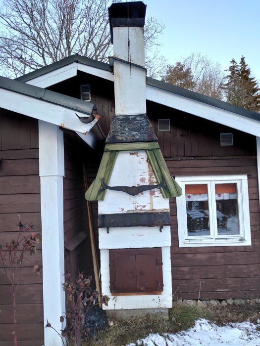 Gammal vitmålad eldstad på en stugas utsida med skorsten och rostig lucka.