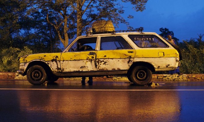 Gammal sliten gul Peugeot 504 taxibil utan bakruta parkerad vid vägkanten om kvällen.