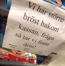 Textskylt med budskapet: "Vi har större bröst bakom kassan, fråga så tar vi fram dem!" ovanpå varor i butik.