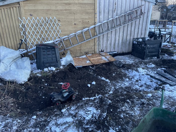 Trädgårdsområde med snötäckta markarbeten, trädgårdsredskap och en fallen stege vid staket.