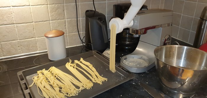 Hemmagjord spagetti pressas från en pastamaskin på ett köksbord med köksredskap.