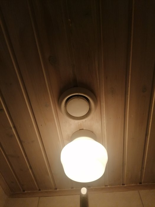 Frånluftsdon bredvid en taklampa på ett träpaneltak i en ombyggd garderob.
