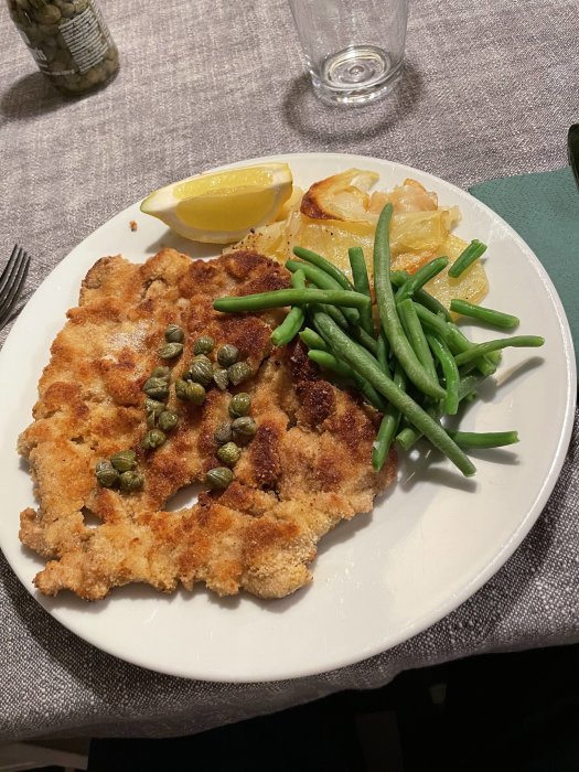 Panerad schnitzel med kapris, citronklyfta, gröna bönor och potatis på en vit tallrik.