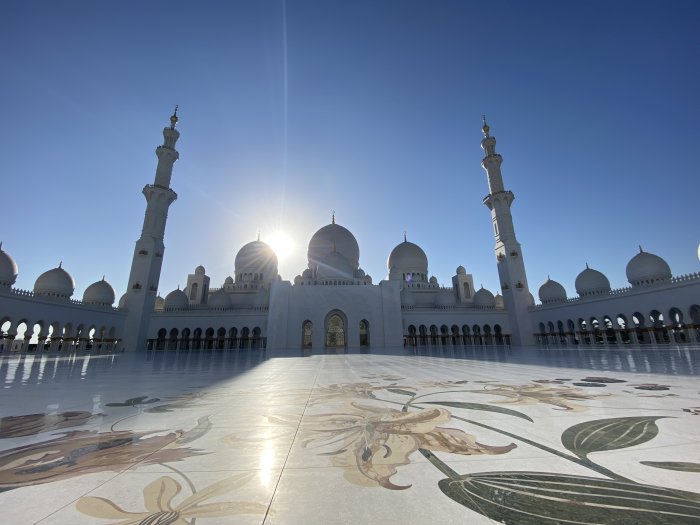 Solnedgång bakom en stor vit moské med minareter och utsmyckade golvplattor.