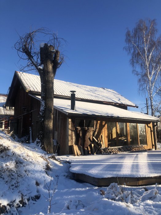 Ett trähus med snötäckt tak och en synlig skorsten mot en klarblå himmel, omgiven av vinterlandskap.