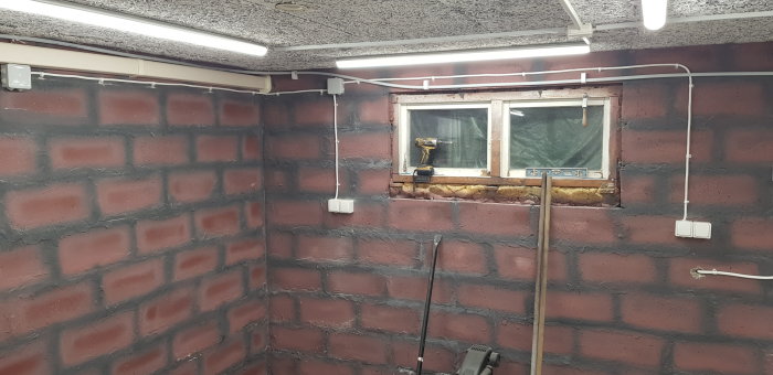 Ett rum under renovering, med synliga murade väggar och installerade elledningar samt vägguttag på ena väggen, intill ett fönster.