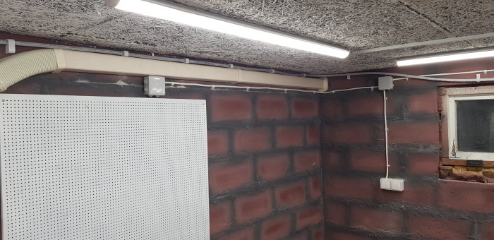 Hörn av verkstad med lysrörsarmatur, klamrad ledning och nyinstallerade vägguttag nära fönster.