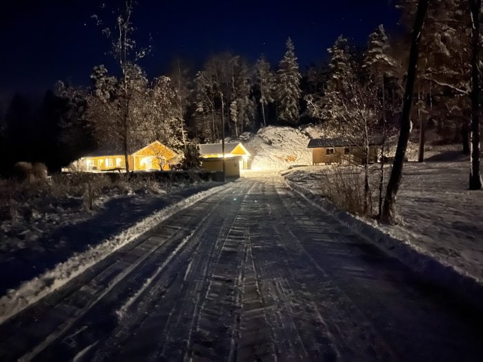 Snötäckt väg plogad av ett gammalt fordon med upplyst hus i bakgrunden på natten.