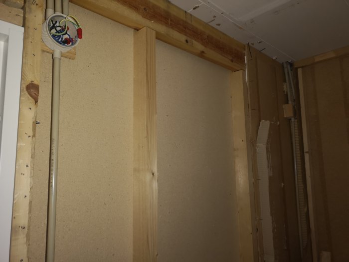 Hörn av en vägg med träreglar och fiberboard, eldosor och synliga elrör, samt indikation på äldre konstruktion bakom.