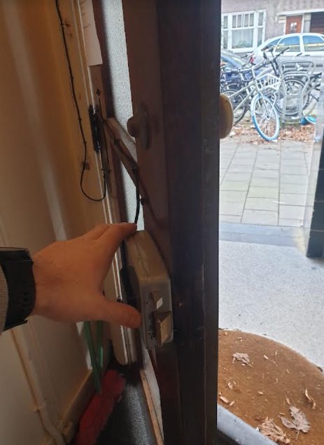 Hand pekar på ett gammalt lås i en trappuppgångsdörr med synliga ledningar och utsikt till cyklar utanför.