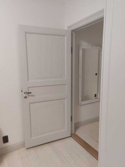 Nyrestaurerad vit dörr öppen mot ett rum med trägolv och spegel.