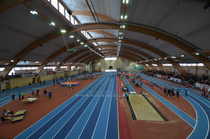Inomhusidrottshall med löparbanor, hoppgruber och atletiska aktiviteter under pågående träningspass.