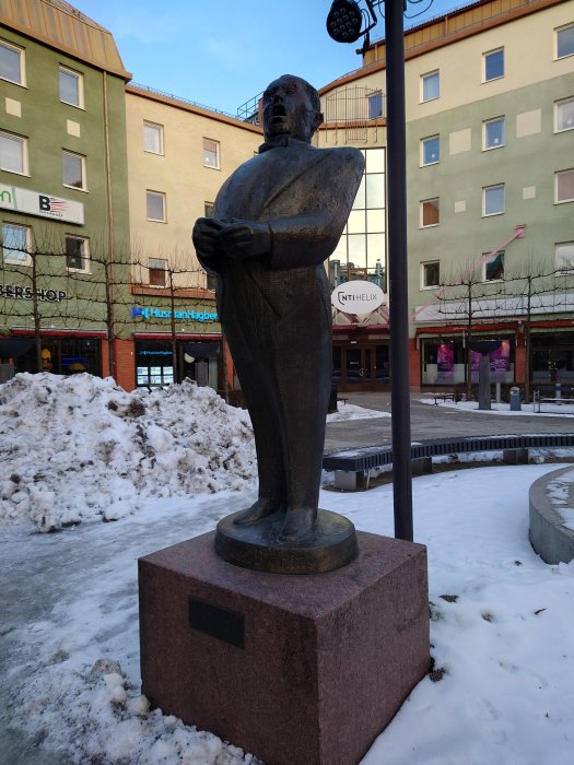 Staty av en man i överkroppen med armar korsade framför snöig bakgrund och stadsmiljö.