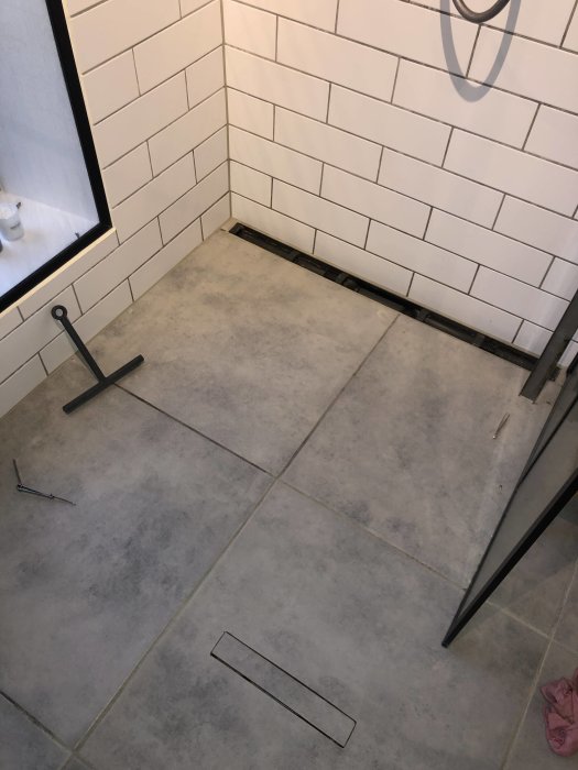 Ett badrumsgolv med två golvbrunnar och vita väggkakel, en golvbrunn nära duschglas.