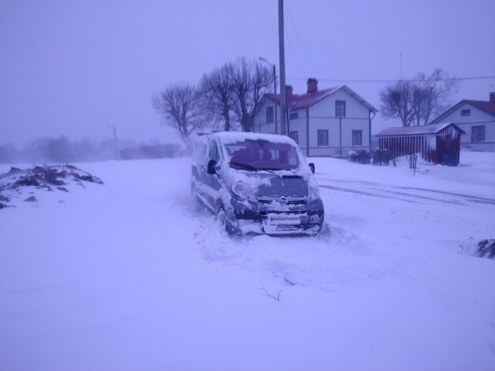 Bil fastkörd i snödriva på en bostadsdrivväg med hus i bakgrunden under vinterförhållanden.