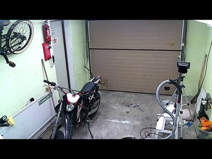 Övervakningskamera bild av ett garage med en motorcykel, cykel, dörr och rörelsedetektor i svagt ljus.