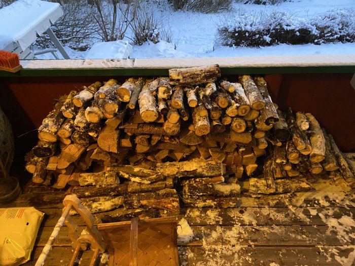 Vedstapel framför röd vägg och snötäckt trädgård i skymningen.