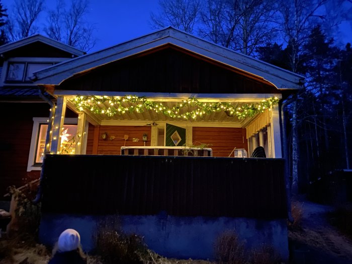 Träveranda på ett rött hus med takbelyst av slingrande ljusslingor i skymningen.