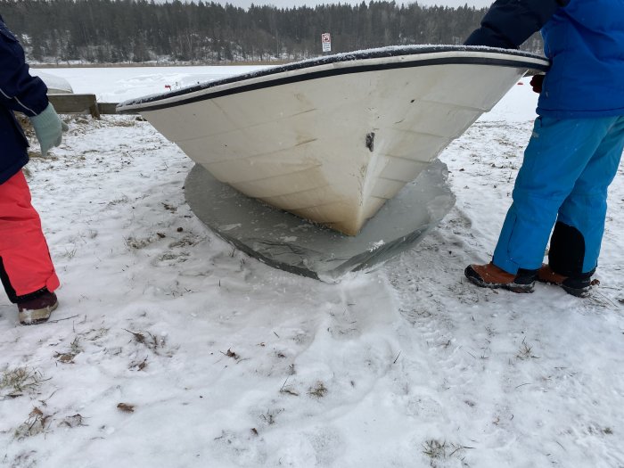 Båt fastfrusen i isen omgiven av snö med två personer i vinterkläder som tittar på.