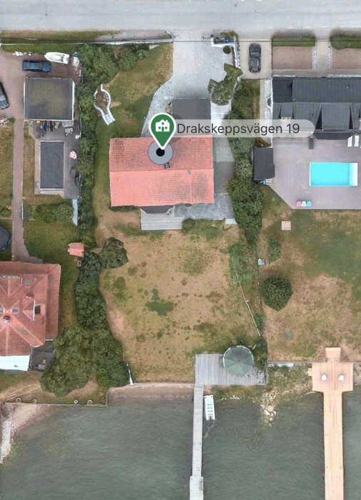 Flygbild över ett fastighet nära vatten med gräsbevuxen yta och en markerad plats, Drakskeppsvägen 19.