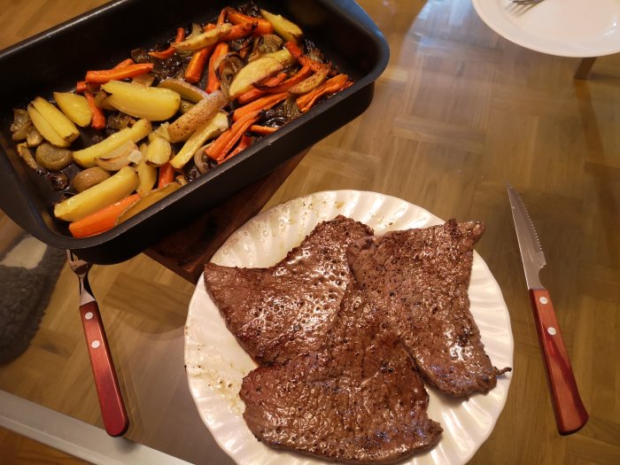 Grillade biffar på en tallrik och rostade rotgrönsaker i en ugnsform på ett träbord.