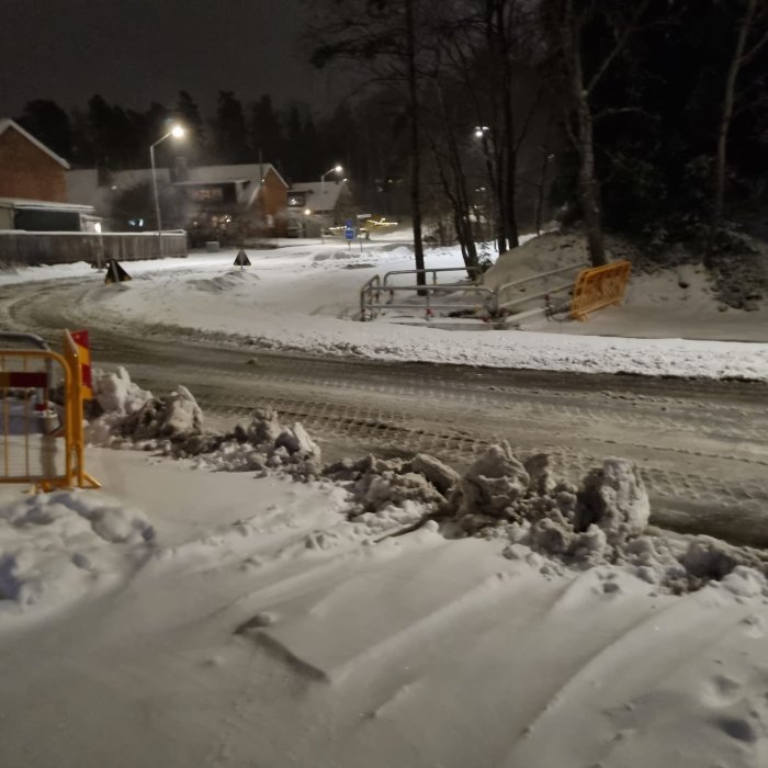 Snöklädd väg med en hög av plogad snö framför ett hus på kvällen.