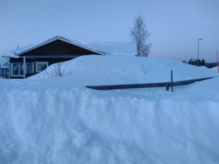 Hus delvis dolt av snötäcket med staket som sticker upp ur snön, vilket ger insynsskydd.