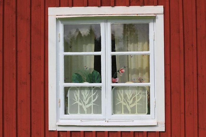 Vitkarmat fönster med spröjs och gardiner mot röd trävägg, små föremål synliga på fönsterbrädan.