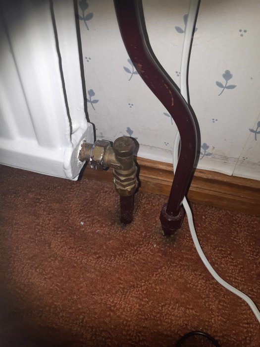 En radiatorventil och rörkoppling vid en vägg med tapet och en brun heltäckningsmatta.