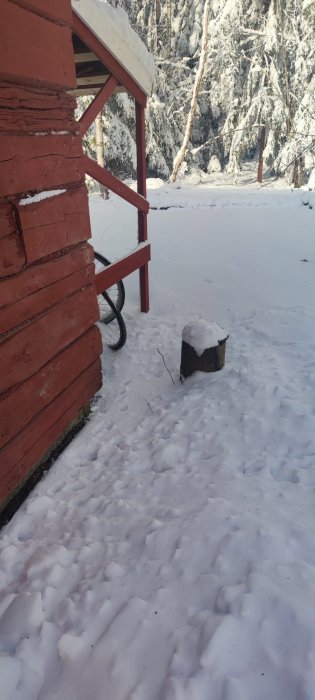 Täckt spolbrunn i snö nära röd timmerstuga med snötäckt tak och snöig skog i bakgrunden.