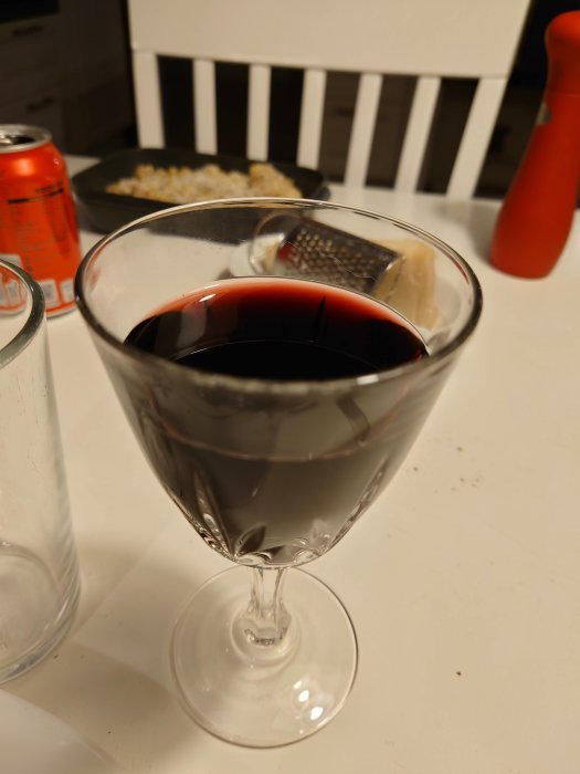 Ett glas rött vin framför en kastrull med mat och en osthyvel på bordet.