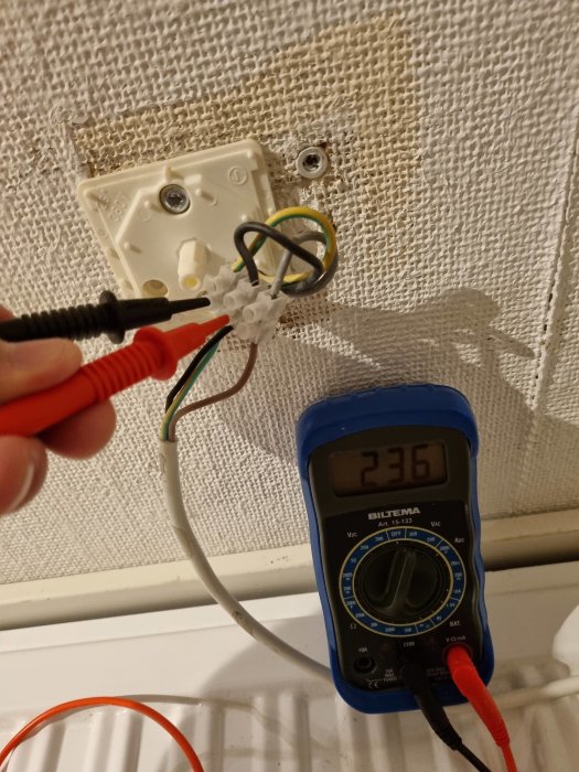 Multimeter visar 236V mätt på kablar vid en väggmonterad elanslutning, bakgrund av texturerad tapet.