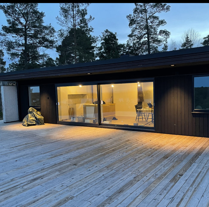 Stor trädäck vid hus med skogsbakgrund och stor glasvägg mot havsutsikt vid skymningen.