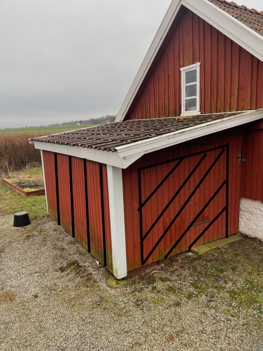 Röd träbyggnad med slitet tegeltak och vit fönsterram, växande mossa längs väggens botten.