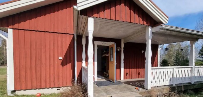 Husets rödmålade gavel med vit veranda och öppen entrédörr under en klarblå himmel.