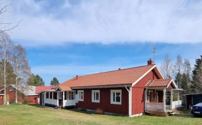 Långsida av rött hus med vita knutar och veranda under en klarblå himmel.