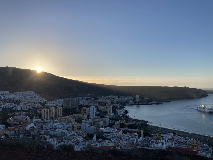 Solnedgång över en kuststad med byggnader, berg i bakgrunden och en bukt i förgrunden.