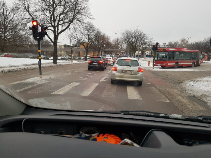 Vy från förarsätet vid trafikljus med bilar och buss, snöfläckar och vägmarkeringar på asfalten.