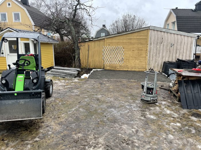 Blöt bakgård med smältande snö, byggmaterial, och kompaktlastare vid gult hus.