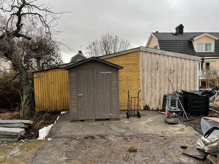 Blöt trädgård med smältande snö, en bod och verktyg mot en molnig himmel.