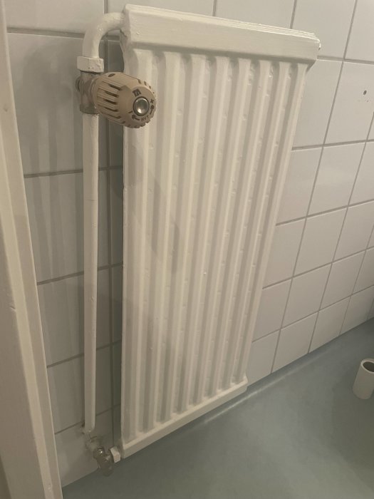 Vattenburet radiatorselement i ett badrum, monterat vid en kaklad vägg med regleringsventil och anslutningsrör.