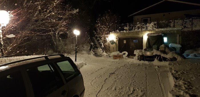 Bil parkerad på snöig uppfart framför garage och hus med tända utomhuslampor i mörkret.