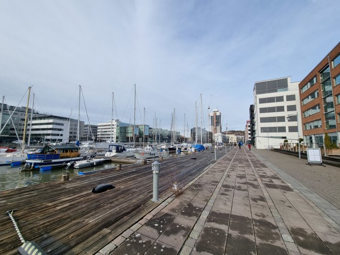 Träbrygga med förtöjda segelbåtar vid moderna byggnader och promenerande personer.
