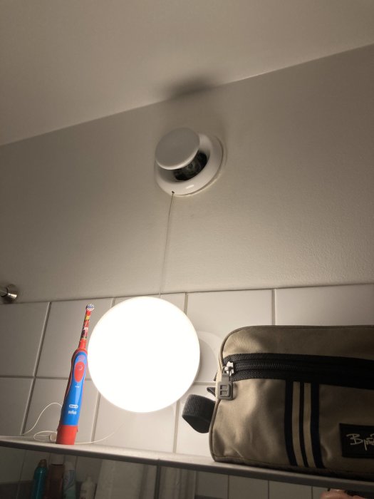 Badrumstak med ventilationsuttag, hyllkant med tandborste och necessär, spegelbelysning i förgrunden.