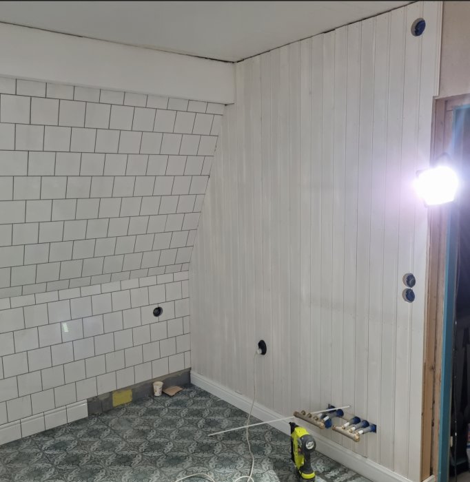 Renoverat badrum med vita kakelväggar, pärlspont och omonterade VVS-installationer på en trävägg.